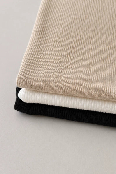 Close-up detail shot of folded ribbed knit fabrics in husk beige, white, and black, showcasing the fine organic cotton and hemp jersey texture.