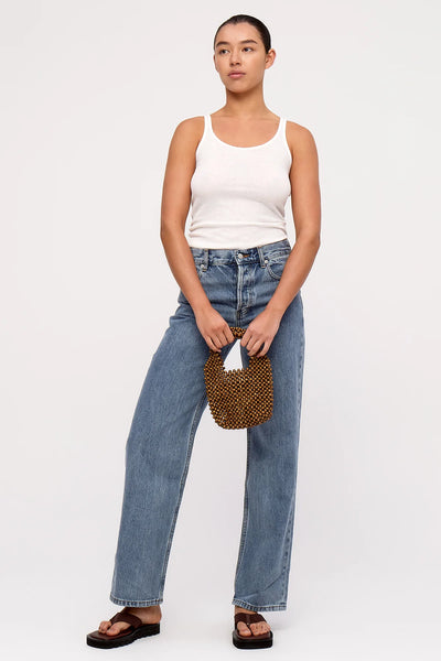 A full-length studio shot of a model dressed in high-waisted wide-leg denim and a white tank top, holding the walnut beaded bag at her side to display its casual, everyday resortwear appeal.