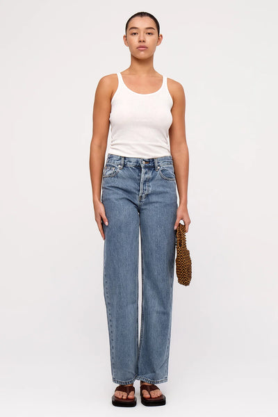 A full-body studio image of a model holding the small walnut wooden handbag with both hands, highlighting how the minimalist silhouette complements a relaxed, modern casual outfit.