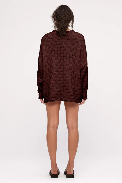 A direct full-length back studio view of the deep brown cotton knit jumper on a model, highlighting the uniform structural continuity of the large-scale weave pattern across the shoulders and back.
