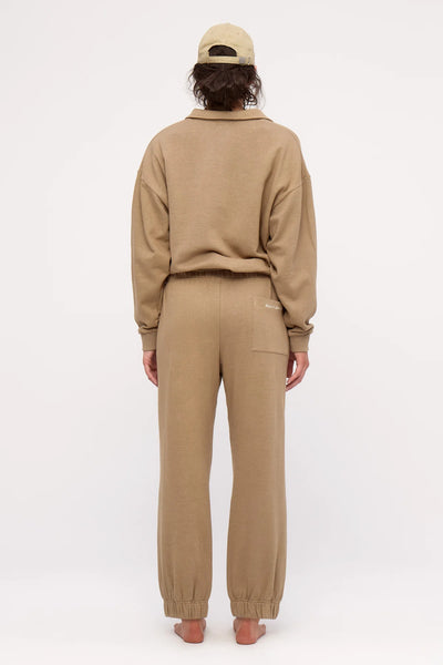 A full-length studio shot from behind, showing a model in a monochrome olive lounge set to highlight the sweatpants' right-side square rear patch pocket and small white embroidered brand logo detail.