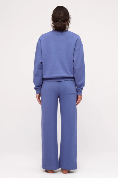 A full-length studio rear view of a model showcasing a matching blue monochrome tracksuit set, highlighting the clean back silhouette of the relaxed, long-sleeve fleece sweatshirt paired with wide-leg track pants.