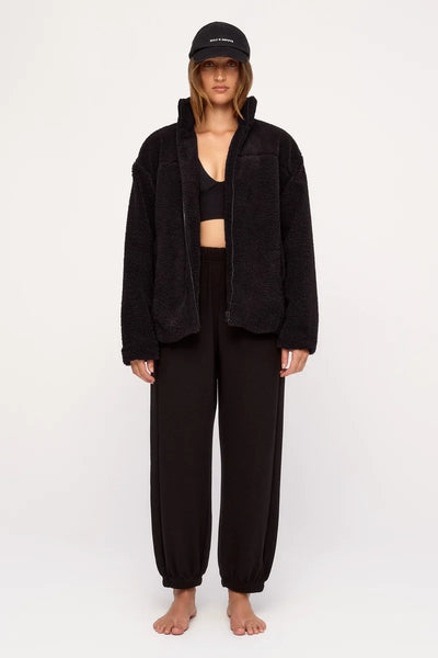 Full-length model shot highlighting the open Zulu & Zephyr Black Sherpa Jacket styled casually with hands in side seam pockets, paired with black shorts and a black cap.