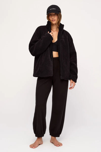 Full-length front view of a model wearing the relaxed Zulu & Zephyr Black Sherpa Jacket unzipped over a black sports crop top and matching track pants, completed with a black logo cap.