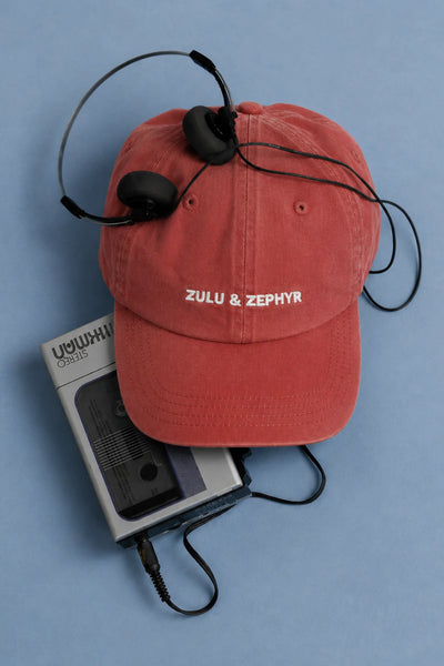 Flat lay studio arrangement of the red pre-washed cotton twill Zulu & Zephyr Canvas Cap resting on a cassette walkman with headphones on a light blue backdrop.