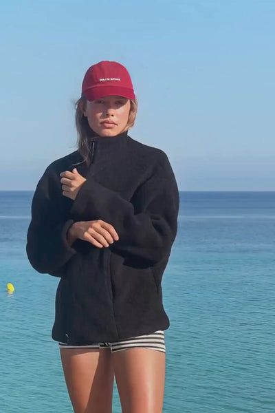 Outdoor lifestyle shot of a model wearing the Zulu & Zephyr Black Sherpa Jacket zipped to the neck with a red baseball cap against an ocean background.