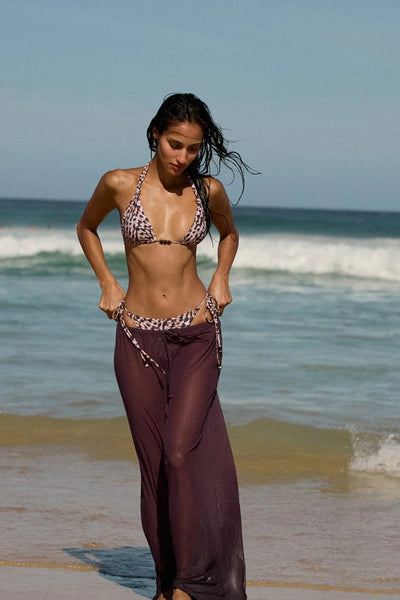 Outdoor beach lifestyle front shot of a model walking near the surf wearing the printed triangle bikini top and matching bottoms paired with a sheer fig purple cover-up skirt.