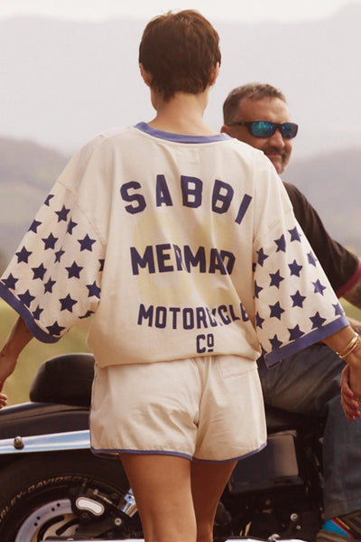 Rear view of a model wearing the cream SABBI Very Oversized Dream Big Brenda Tee, displaying the blue collegiate back text "SABBI MERMAID MOTORCYCLE CO" and star patterns on the wide sleeves.
