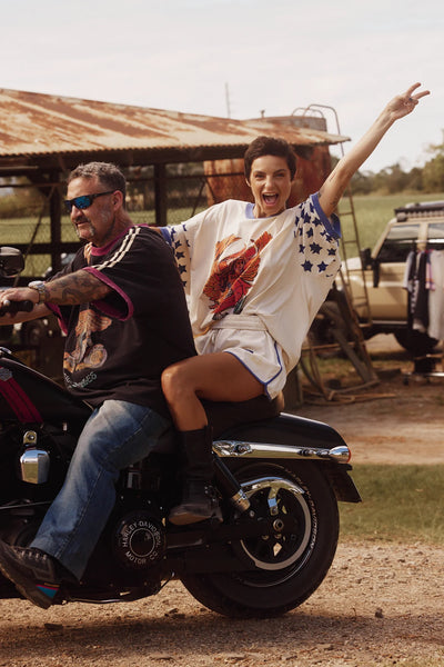 Action shot of a female model giving a peace sign while riding passenger on a motorcycle, wearing the cream SABBI Very Oversized Dream Big Brenda Tee featuring a central winged-mermaid graphic and star-spangled sleeves.