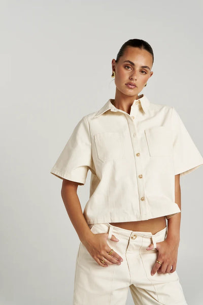 Mid-shot portrait of a model posing in the cropped Summi Summi Carpenter SS Shirt in Ecru, styled with matching Ecru trousers and gold jewellery.
