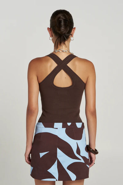 Studio back view of a model highlighting the symmetrical cross-over back strap design and a rounded keyhole cutout on the ribbed knit Espresso Halle Tank, tucked into a brown and blue abstract print mini skirt.