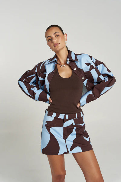 Studio style campaign shot featuring a model posing with hands on her hips, styling the figure-hugging Summi Summi Halle Tank in Espresso under an unbuttoned blue and brown abstract print jacket with a matching mini skirt.