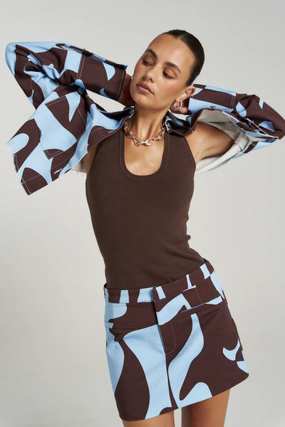 A dynamic studio shot of a model with her hands behind her head, showcasing the scoop neckline of the brown ribbed Halle Tank top paired with an unbuttoned blue and brown patterned jacket, a silver link necklace, and a matching print mini skirt.