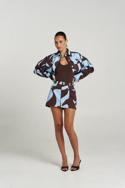 Full-length studio style shot of a model wearing the tight-fitting organic cotton Halle Tank in Espresso, layered under a bold blue and brown abstract print jacket and matching mini skirt, finished with brown kitten heel sandals.