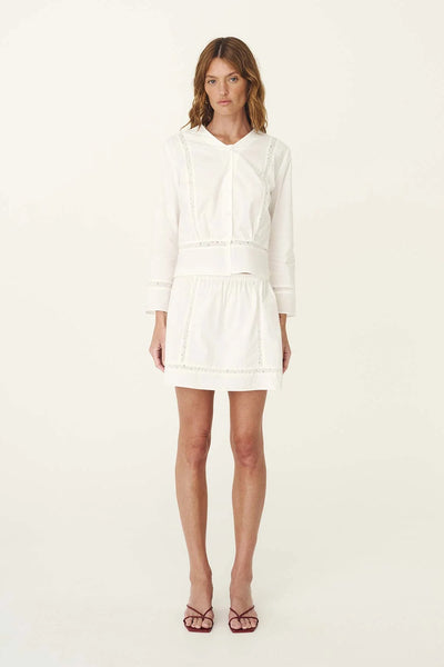 Studio front full-length view of a model wearing the white Suri lace mini skirt and matching long-sleeve blouse with red heeled sandals.