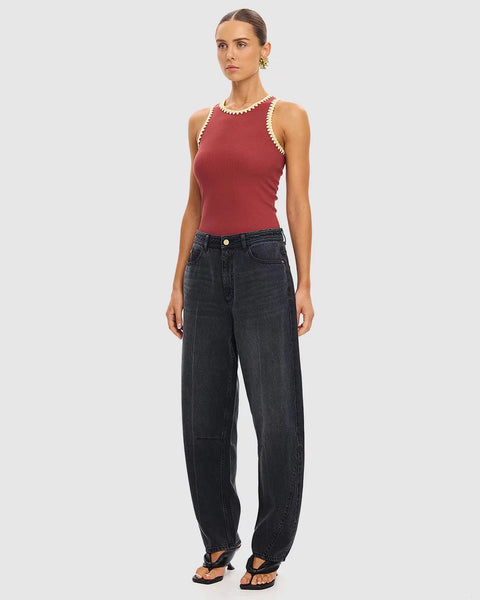 A three-quarter angle studio shot of a model posing in the brick red Kivari Tallulah Spliced Tank. The portrait captures the premium soft ribbed knit fabrication, the artisanal cream crochet details, and the overall fitted silhouette of the tank top paired with wide-leg charcoal jeans.