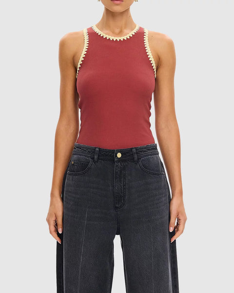 A waist-up studio portrait of a model modeling the Kivari Tallulah Spliced Tank in Brick. The shot clearly showcases the high-neck design, the distinct beige crochet trim contrasts against the rust fabric, and the close-to-body fit, styled with statement gold earrings and washed black denim.