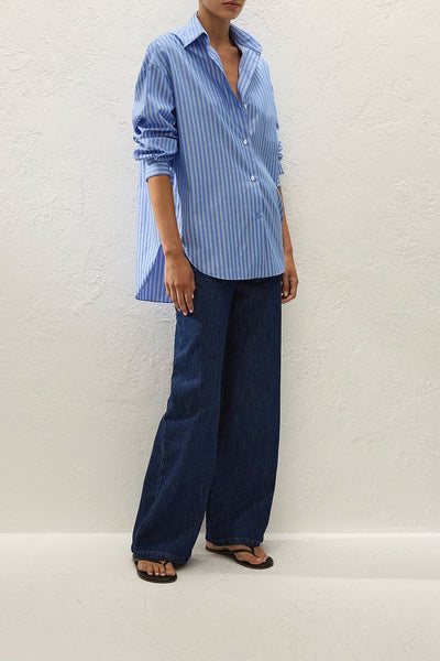Full-length three-quarter shot of a model styling the relaxed, oversized Faithfull The Brand En Vau Shirt in Joao Stripe Azulejo Blue with dark denim trousers and flat sandals.