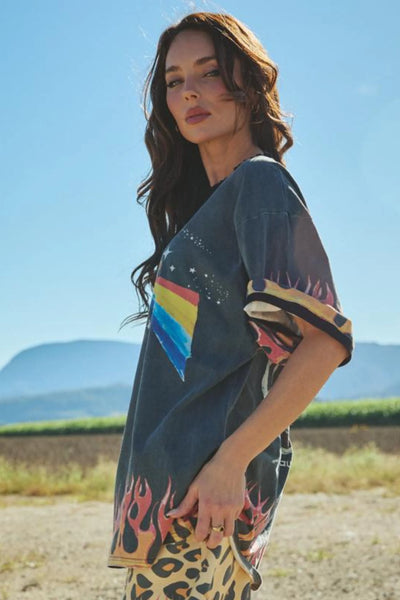 A side-angle photograph isolating the washed black cotton jersey on a model, emphasizing the dramatically loose fit, oversized armholes, and the vibrant orange and yellow statement flame graphics that wrap around the side panel.