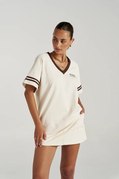 Upper-body studio view of a model styling the Summi Summi Carpenter Skirt in Ecru with a matching cream V-neck tee featuring brown contrast trim.