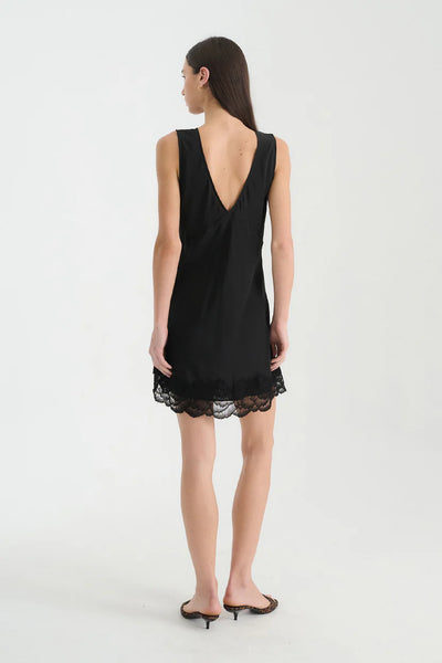 Rear view of a model styled in low heels wearing the ROWIE Annie Silk Mini Dress in Noir, displaying the low V-back cut and sheer lace-trimmed straight hemline.
