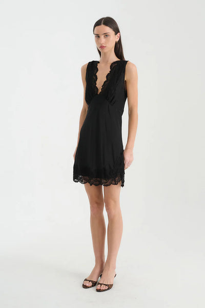Full-length studio portrait of a model standing straight in the ROWIE Annie Silk Mini Dress in Noir, featuring a scalloped V-neck, fitted empire bust, and lace-trimmed hem.