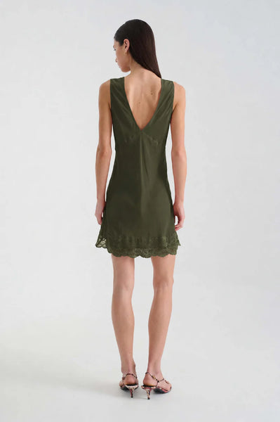 Full-length back view of a dark-haired model wearing the ROWIE Annie Silk Mini Dress in Olive styled with low heels, featuring a low V-back cut and fluid bias-cut silk drape.