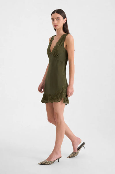 Three-quarter side profile of a model walking in the ROWIE Annie Silk Mini Dress in Olive and snakeskin mules, illustrating the mini length, asymmetrical side split, and lace detailing.