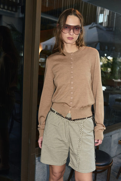 Campaign image of a model outdoors wearing the ROWIE Maeve Merino Spring Cardigan in Fawn Marle styled with plaid shorts and tinted sunglasses.