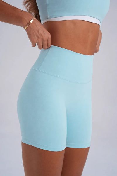 Side angle studio shot of a model adjusting the seamless tapered high-rise waistband of ice blue activewear bike shorts.