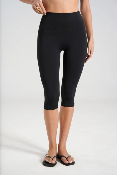 Studio front view focusing from the waist down on a model wearing high-waisted black 3/4 length capri leggings and flat flip-flops.