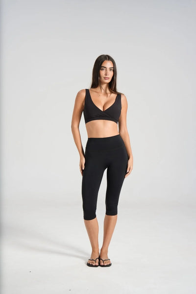 Full-length studio front view of a brunette model wearing flat flip-flops, a black V-neck sports bra, and high-waisted black capri leggings with a seamless front panel.