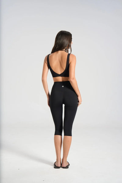 Full-length studio rear view of a model wearing flat thong sandals, a black cross-back sports bra, and high-waisted 3/4 length black capri leggings.