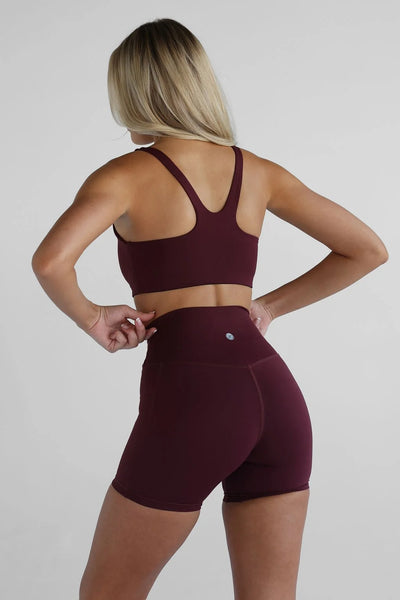 Studio rear view of a blonde model with hands on her hips, wearing a deep maroon activewear set including high-waisted 6-inch bike shorts and a matching racerback sports bra.
