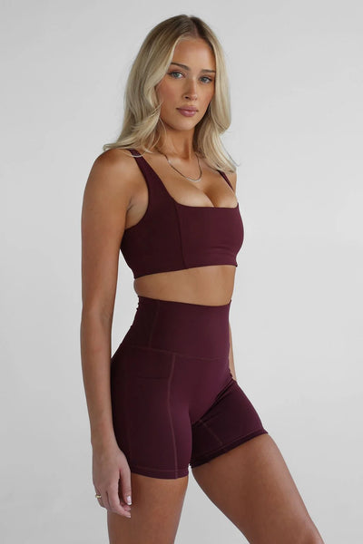 Studio three-quarter front view of a blonde model wearing high-waisted dark cherry cola bike shorts with side drop-in pockets and a matching square-neck sports bra.