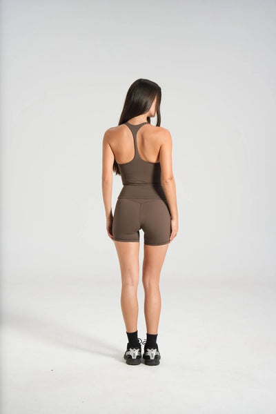 Full-length studio rear view of a model with long dark hair wearing black athletic socks, sneakers, and a dark chocolate brown slim racerback tank top paired with matching high-waisted shorts.