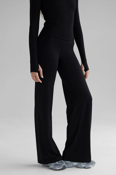 A three-quarter studio profile view of a model wearing the black LEELO Sculpt Wide Leg Pants and a matching black long-sleeve top. The photograph highlights the relaxed drape and elegant cut of the legs, paired with grey-blue athletic sneakers against a plain grey backdrop.