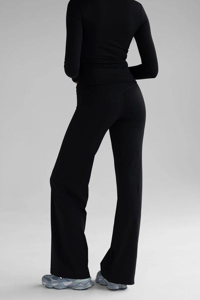 An angled rear studio view of a model wearing the LEELO Sculpt Wide Leg Pants in black. The pants are styled as a monochrome athleisure set with a matching black long-sleeve performance top, highlighting the seamless integration of the high-rise pants.
