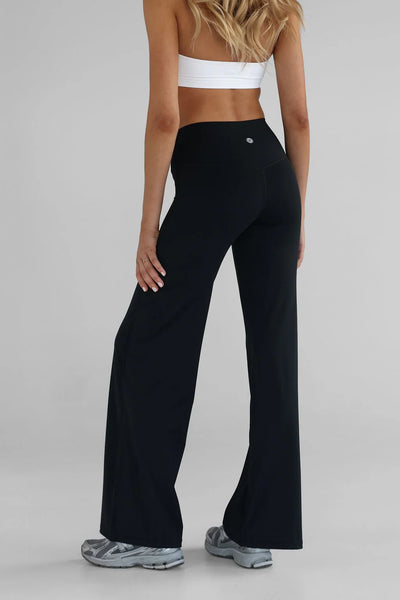 Rear studio view from the waist down of a model wearing the black LEELO Sculpt Wide Leg Pants. The shot emphasizes the smooth shape of the brushed nylon-spandex blend, the V-shaped back seam lines, and a small silver silicone logo centered on the high waistband.