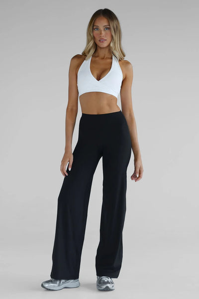Full-length studio shot of a model showcasing the LEELO Sculpt Wide Leg Pants in black. The pants feature a long 33-inch inseam length and a fluid, wide-leg silhouette, paired with a contrasting white wrap-front halter neck sports bra and grey athletic sneakers.