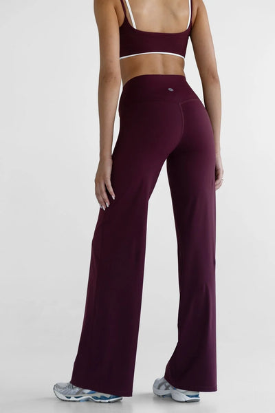 Back three-quarter view of a model wearing the high-waisted Leelo Sculpt Wide Leg Pants in Cherry Cola paired with a matching sports bra and athletic sneakers.