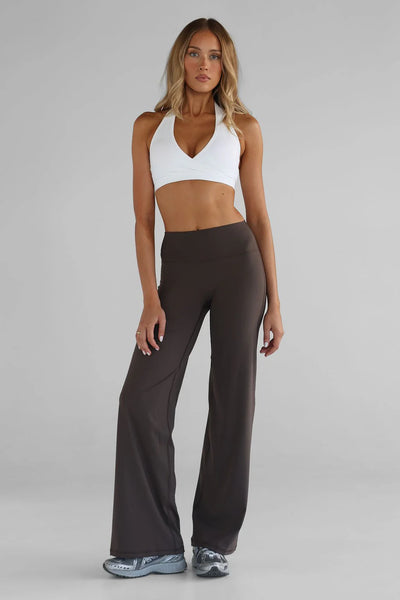 Full-length studio shot of a model wearing the LEELO Tall Sculpt Wide Leg Pants in dark chocolate brown. The pants feature a long 33-inch inseam length and a relaxed leg design, paired with a white halter neck sports bra and athletic sneakers against a neutral grey studio backdrop.