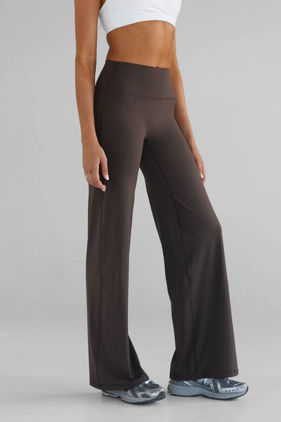 A side profile studio view focusing on the lower body showcasing the LEELO Tall Sculpt Wide Leg Pants in dark chocolate brown. The image emphasizes the supportive high-rise waistband, smooth hip line, and the relaxed drape of the wide-leg cut, styled with grey athletic sneakers.