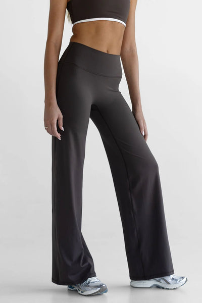 A side profile studio image highlighting the long, fluid silhouette of the dark chocolate LEELO Tall Sculpt Wide Leg Pants. The model is shown from the ribs down, showcasing the clean side lines and durable stitching of the premium nylon-spandex blend activewear fabric.