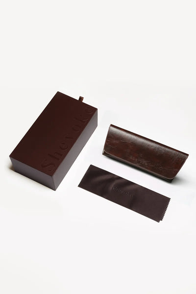 Overhead view of the Shevoke protective packaging set featuring a dark brown branded outer box, a leatherette folding sunglasses case, and a microfiber cleaning cloth.