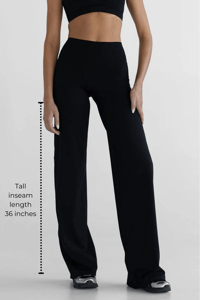 A full-length studio product photograph of a tall model from the front wearing the black Tall Sculpt Wide Leg Pants, paired with a matching black sports bra. The image highlights the smooth, no-front-seam construction, the seamless tapered high waistband providing tummy control, and the relaxed flare of the wide-leg silhouette.
