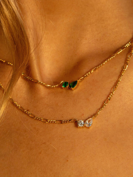 Close-up shot of two layered gold necklaces with emerald-cut and pear-cut green stone pendants glowing in warm sunlight.