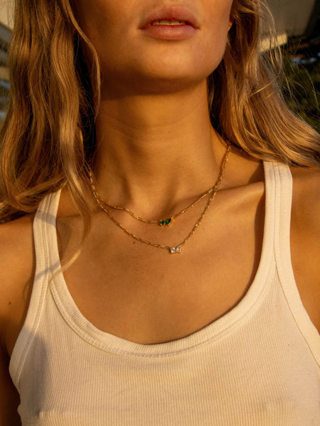 Close-up of a model wearing two layered gold necklaces, including the Twin Flame necklace, against a white tank top under warm sunlight.
