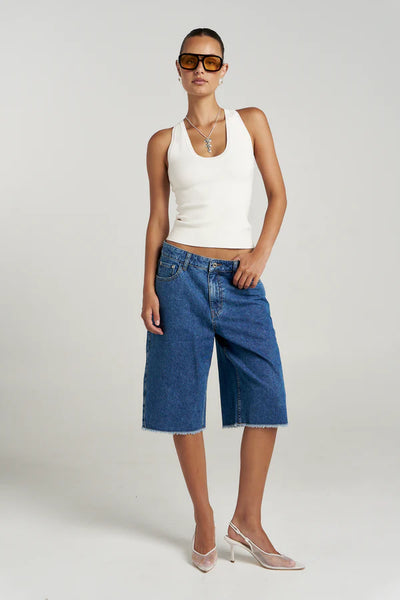 Full-length studio campaign shot showcasing the versatile Vanilla Halle Tank top paired with oversized, raw-hem long blue denim shorts and white slingback kitten heels against a neutral background.