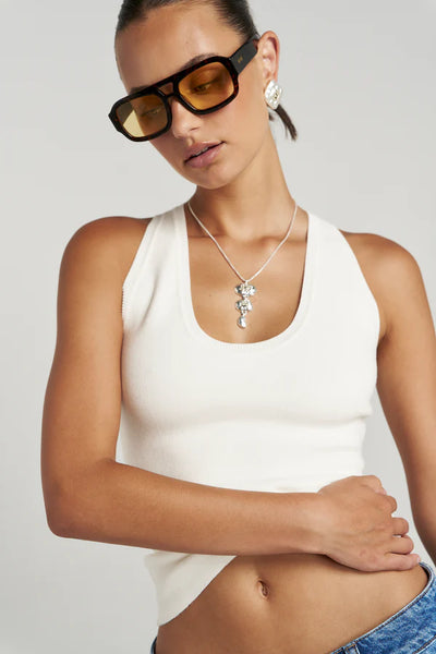 A close-up studio shot of a model wearing the ribbed knit Vanilla Halle Tank, accessorized with a chunky silver statement necklace and tortoiseshell aviator sunglasses with yellow lenses.